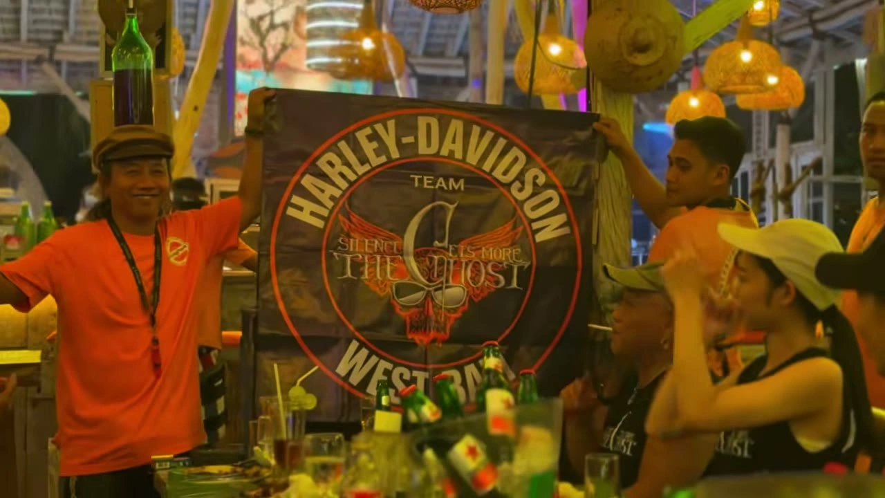 オレンジシャツの男性がTHE GHOSTの看板・正面笑顔｜2026年6月 バリ島ハーレーツーリング — TEAM THE GHOST West Bali